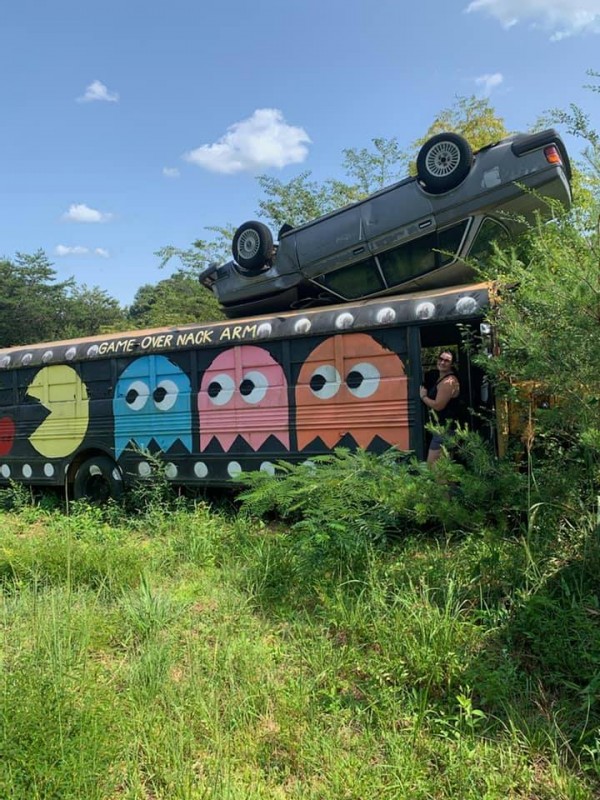 a train car in a field of green grass and a blue sky overhead. There is a pac man design painted on the train with a black background a dot with a pac man and three ghosts (blue, pink, orange) Beth Wiza stands at the right end of the train car, near the orange ghost and points at it, smiling.