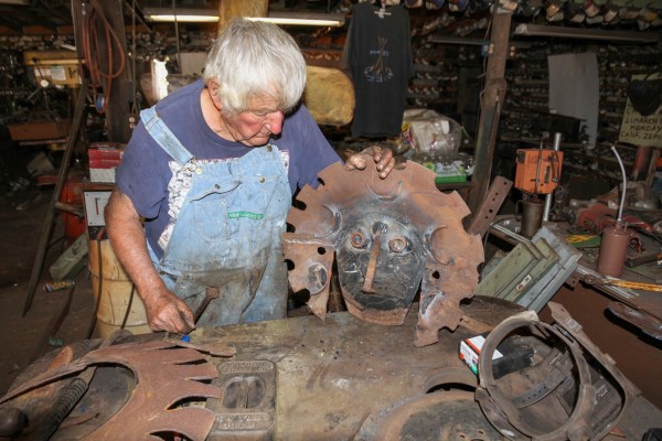 Artist M.T. Liggett is at a table covered in rusted metal scraps and holding a mask-like sculpture made of metal